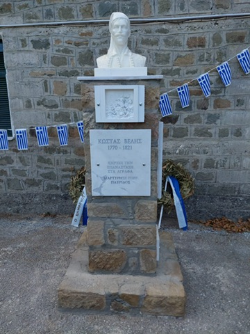 An outdoor memorial: a white stone bust of the Greek revolutionary Kostas Velis, moustached and wearing a slanting cap and traditional vest, crowning a tall square-cut stone pillar. The pillar's front carries a marble plaque reading 'ΚΩΣΤΑΣ ΒΕΛΗΣ 1770 - 1821 / ΚΗΡΥΞΕ ΤΗΝ ΕΠΑΝΑΣΤΑΣΗ ΣΤΑ ΑΓΡΑΦΑ / ΜΑΡΤΥΡΗΣΕ ΥΠΕΡ ΠΑΤΡΙΔΟΣ' — Kostas Velis, 1770 to 1821, he proclaimed the revolution in Agrafa, he gave his life for the fatherland — with a smaller relief-carved plaque of a battle scene above it. Behind, a stone masonry wall is strung with two rows of blue-and-white Greek flag bunting, and two dried olive wreaths with blue-and-white ribbons lean against it either side of the pedestal.