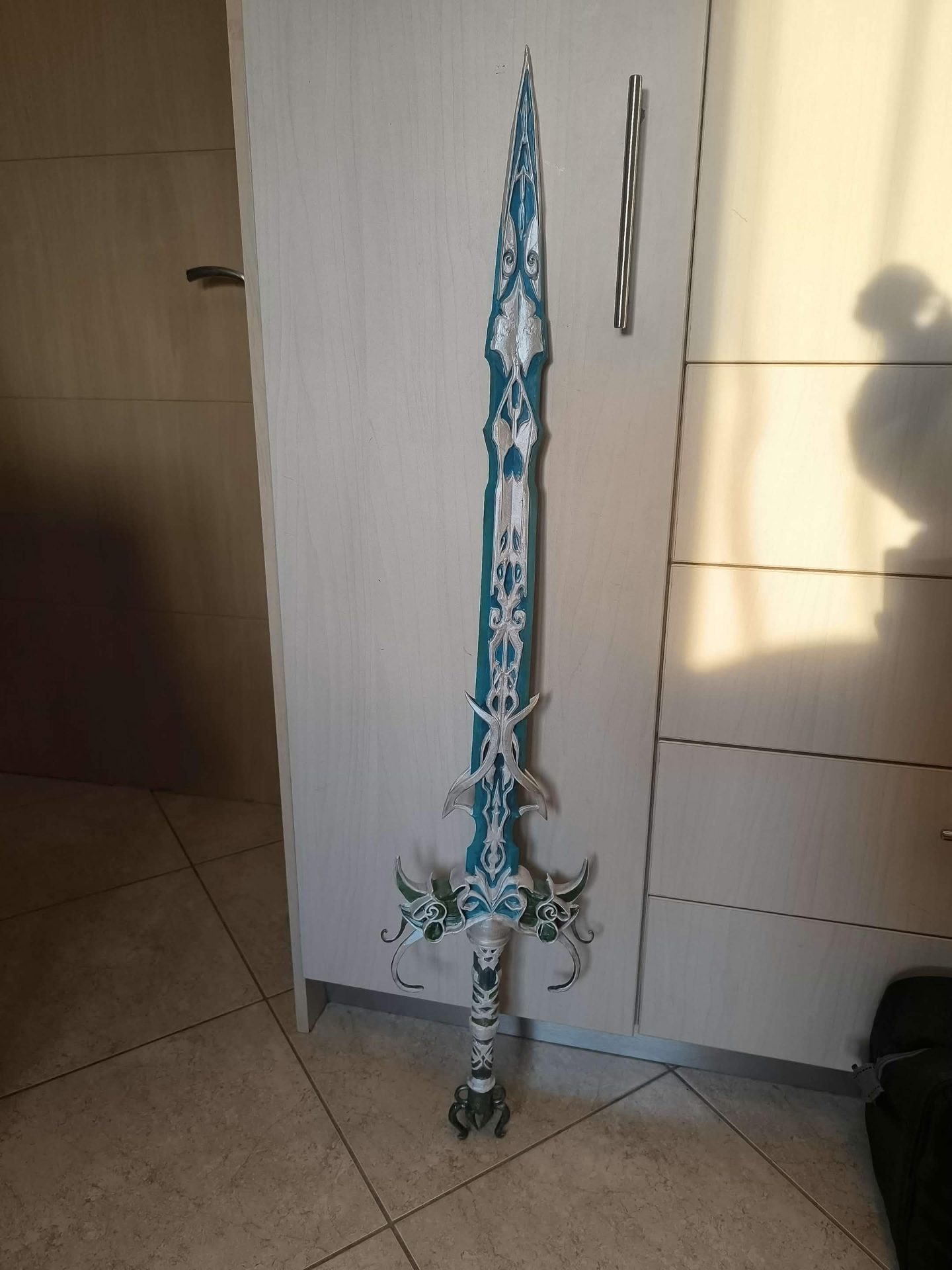 A photograph of the same sword built as a real prop, leaning against a light-wood wardrobe on a tiled floor: metallic silver and white detailing picked out with turquoise paint inlay.